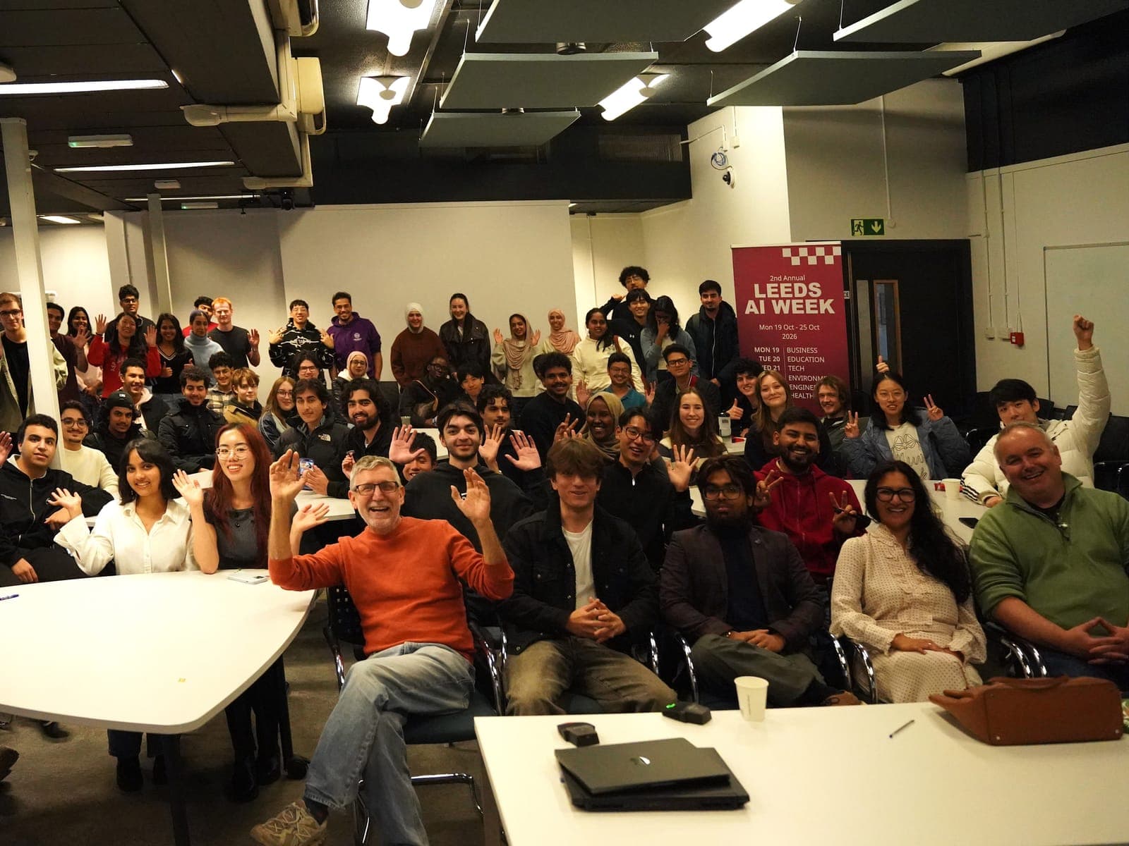 Around sixty hackathon participants waving at the camera in a university teaching room, with a Leeds AI Week banner on the right